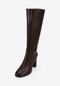 Botas de cuero marrón oscuro, de caña alta, con tacón cuadrado y una pequeña correa decorativa con hebilla por encima del tobillo, sobre un fondo liso.
