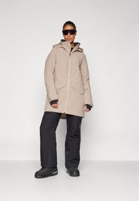 Chaqueta impermeable beige con capucha y cremallera frontal, combinada con pantalones de esquí negros y botas negras resistentes, que presentan materiales texturizados.