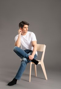Weißes Crew-Hals-T-Shirt, hellblaue Denim-Jeans und schwarze Socken auf einem Holzstuhl. Grauer Hintergrund, sitzende Pose mit übergeschlagenen Beinen.