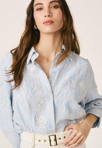 Femme aux longs cheveux bruns portant une chemise bleu clair à boutons avec une broderie florale blanche et une ceinture ivoire à boucle carrée.
