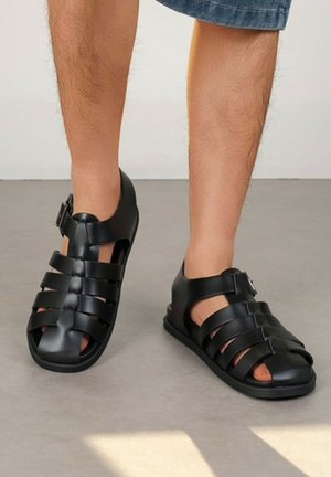 Jambes portant des sandales noires à bouts fermés avec plusieurs sangles horizontales et boucles, debout sur un sol gris en short en jean.