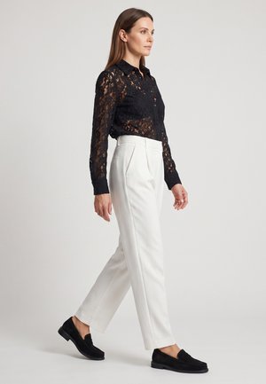 Kvinde går iført en sort blonder langærmet bluse, hvide bukser med høj talje og sorte loafers mod en ensartet grå baggrund.