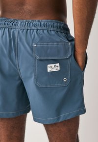 Plave kupaće kratke hlače s elastičnim pojasom, stražnjim džepom sa zakrpom, bijelim prošivom i etiketom s logotipom na kojoj se nalazi prikaz palme.