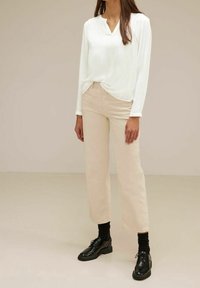 Top bianco a maniche lunghe con scollatura a V e nodo frontale. Pantaloni beige a gamba larga con una texture liscia. Scarpe nere con lacci e suola spessa.