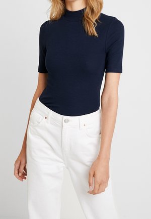 Kobieta ubrana w granatowy krótki rękaw i półgolf, włożony w białe, wysokowysokościowe dżinsy przeciwko jednolitemu tłu.