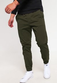 Olijfgroene joggerbroek van lichtgewicht stof met elastische boorden, zijzakken en een taille met trekkoord, gecombineerd met witte sneakers.