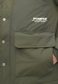 Casaco isolante verde-oliva com um grande bolso frontal, botões de pressão e texto do logótipo branco. Textura do tecido lisa, apresentando um design de corte confortável.