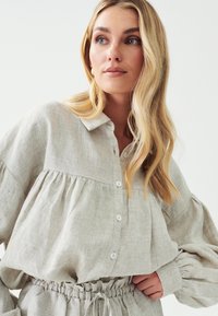 Světle béžová lněná košile s knoflíkovým zapínáním, stojáčkem a nařasenými rukávy. Tkanina má jemnou texturu a uvolněný střih.