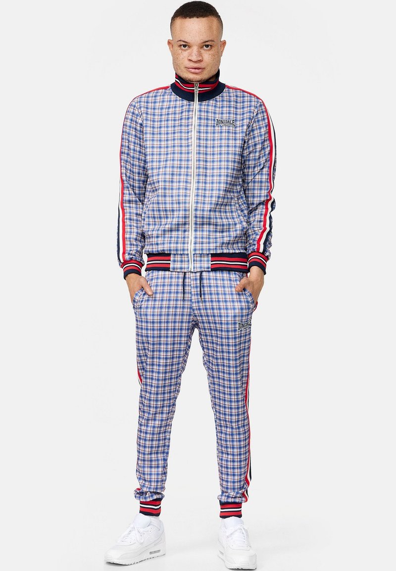 Blauw en wit geblokt trainingspak met rode en zwarte gestreepte accenten. Bevat een zipjack en bijpassende broek, gecombineerd met witte sneakers.