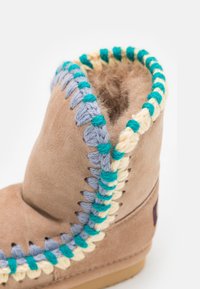 Bota de ante marrón con una parte superior redondeada, que presenta costuras decorativas en hilo azul, verde y crema a lo largo de la abertura y los laterales.