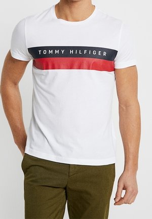 Mężczyzna ubrany w białą koszulkę z krótkim rękawem z granatowo-czerwonymi poziomymi paskami i napisem "Tommy Hilfiger", w połączeniu z oliwkowozielonymi spodniami.