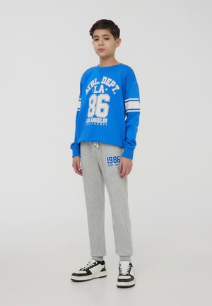 Ragazzo in piedi con pantaloni della tuta grigi e una felpa blu con testo bianco, che indossa scarpe da ginnastica in bianco e nero su uno sfondo neutro.