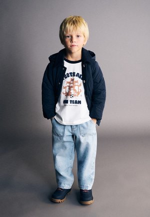 Băiat blond în jachetă bleumarin, tricou alb cu animale de desene animate și text, blugi deschiși la culoare și pantofi închiși, stând cu mâinile în buzunare.