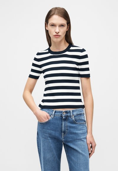 Γυναίκα με κοντομάνικη μπλούζα με οριζόντιες ασπρόμαυρες ρίγες και μπλε ψηλόμεσα τζιν, με το χέρι στην τσέπη, στέκεται μπροστά σε μια απλή φόντο.