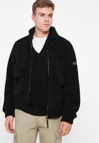 Ung man iklädd en svart fleecejacka över en svart v-ringad tröja och vit skjorta, kombinerad med beige cargobyxor, stående mot en vit bakgrund.