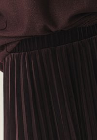 Bordeaux geplooide rok van een gestructureerde, fluwelen stof. Beschikt over een gerimpelde tailleband en verticale plooien voor een gestructureerd uiterlijk.