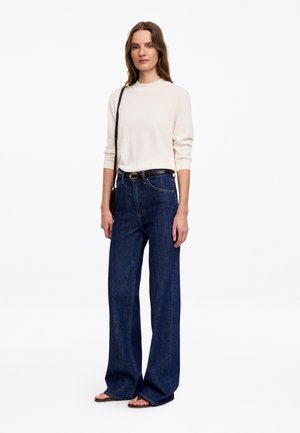 Femme debout portant un haut crème à manches longues, un jean bleu foncé à jambes larges, une ceinture noire, une sangle de sac marron et des sandales ouvertes sur fond blanc.