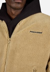 Jachetă din fleece bej cu o textură moale, având un fermoar frontal și logo-ul "PEGADOR" în negru pe piept. Strat inferior cu glugă neagră.
