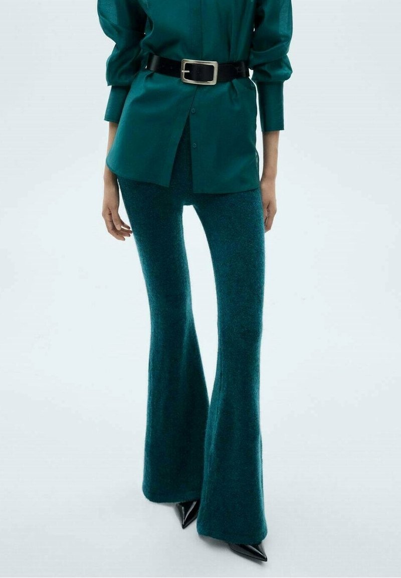 Pantaloni a campana color turchese con una texture morbida, abbinati a una camicia button-up verde scuro e una cintura nera con una fibbia rettangolare in metallo.