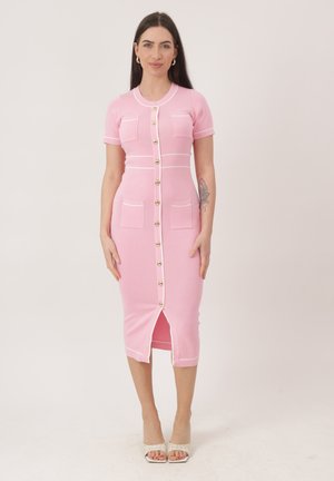 Mujer de pie, vestida con un vestido midi ajustado rosa con botones dorados, mangas cortas, bolsillos delanteros y ribete blanco, combinado con tacones blancos.