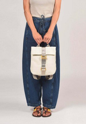 Persona con ropa informal sosteniendo una mochila color crema con correa gris y hebilla amarilla, vistiendo pantalones vaqueros holgados y sandalias.