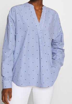 Frau trägt eine blau-weiß gestreifte Bluse mit schwarzen Punkten, langen Ärmeln, V-Ausschnitt und weiße Hose, eine Hand in der Tasche.