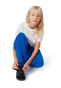 Witte t-shirt met een roze hart graphic, gecombineerd met blauwe geribbelde broek. Persoon die zwarte, gestructureerde sneakers met kleurrijke veters vastbindt.