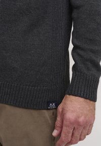 Mörkgrå stickad tröja med ribbade ärmslut och nederkant, med en liten etikett på sidan. Materialet verkar vara mjukt och texturerat.