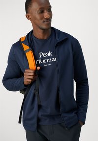 Navy zip-up takki navy t-paita, jossa on valkoista tekstiä. Malli pitää mustaa laukkua, jossa on oranssi hihna. Kangas näyttää sileältä ja mukautuu hyvin.