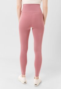 Hoge taille roze leggings gemaakt van rekbaar materiaal; gladde textuur met een aansluitend ontwerp. Een paar lichte schoenen zichtbaar bij de voeten.