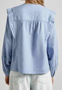 Hellblaue Bluse mit gerafftem Rücken, Spitzenakzenten an Schultern und Ärmeln sowie Ballonärmeln. Weiches Baumwollmaterial, lockere Passform.