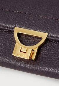 Purple textured leather handbag flap with a gold metallic clasp and branding detail. Features clean lines and subtle stitching accents.