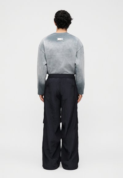 Άνδρας με σγουρά σκουρόχρωμα μαλλιά που στέκεται με την πλάτη, φοράει γκρι μπλούζα με μακριά μανίκια και άνετο μαύρο παντελόνι σε απλό φόντο.