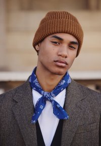 Bruine geribde gebreide muts, blauwe paisley sjaal gestrikt om de hals, visgraatblazer over wit overhemd, close-up van een jonge mannelijke model.