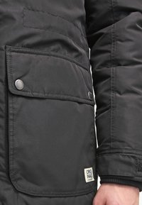 Svart isolerad jacka med sidoficka, tryckknappsstängning och en tygetikett med texten "ORIGINAL." Slät textur och puffy design.