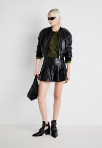 Giacca bomber in pelle nera, maglione con stampa leopardata verde, gonna plissettata in ecopelle nera, stivali neri alla caviglia con cinturini e borsa a tracolla nera.