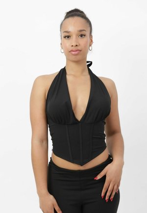 Mujer joven con el cabello recogido en un moño, vistiendo un top halter negro y pantalones negros, de pie con una mano en la cadera contra un fondo blanco.