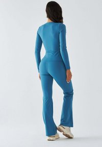 Niebieski, długi rękaw strój treningowy z dopasowaną górą i rozszerzanymi spodniami wykonanymi z gładkiego, elastycznego materiału, uzupełniony neutralnymi butami sportowymi.