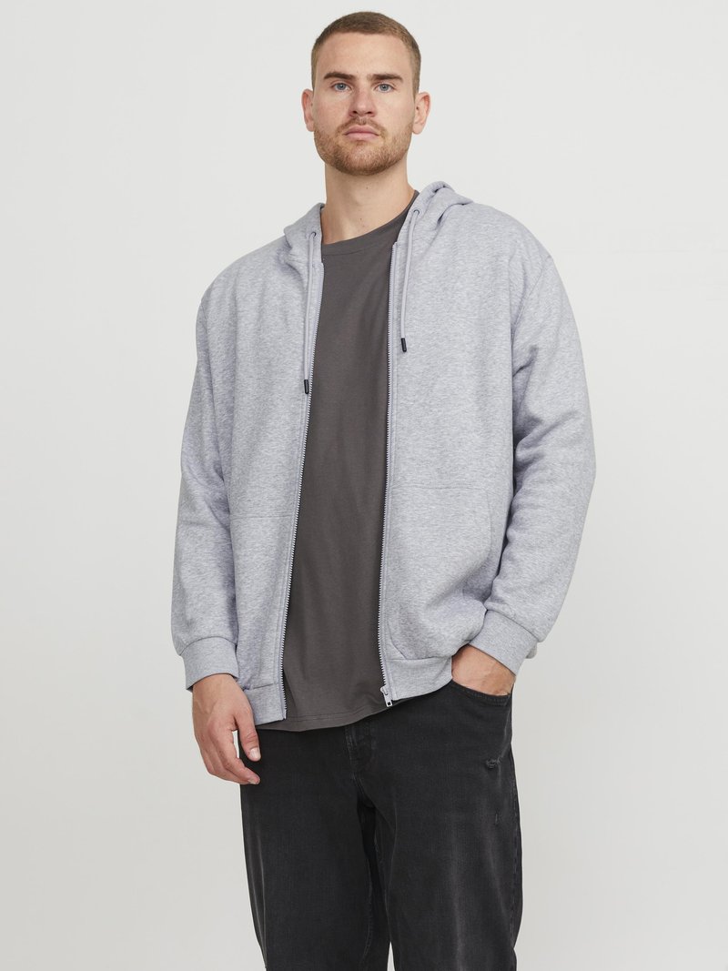 Grå hettegenser med glidelås laget av et mykt stoff. Har ribbestrikkede mansjetter og to sidelommer, brukt over en mørk t-skjorte og sorte jeans.