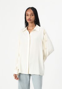 Blusa color crema con bottoni sul davanti, maniche lunghe a sbuffo e dettagli con bottoni sulle braccia. Realizzata in un tessuto liscio.