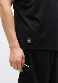 T-shirt de sport noir en tissu texturé, avec des accents rouges sur les côtés et un écusson blanc avec logo sur l'ourlet. Le modèle tient la couture latérale.