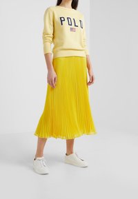 Donna che indossa un felpa "POLO" giallo pallido, una gonna midi plissettata gialla brillante e sneakers bianche con lacci, in piedi su un pavimento bianco.