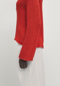 Pull rouge en maille avec une coupe ample et des manches longues. La texture est ouverte et aérée, avec un ourlet droit. Associé à un pantalon blanc.