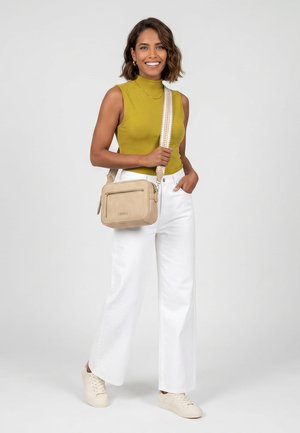 Mujer sonriente con top verde sin mangas, pantalones blancos de pierna ancha, zapatillas blancas, llevando un bolso cruzado beige con correa estampada sobre fondo gris.