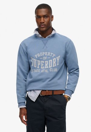 Blauwe sweatshirt met een ronde hals, "Superdry" logo in het wit, geribde mouwen, gelaagd over een lichtgestreept shirt. Donkere broek en een bruine riem.