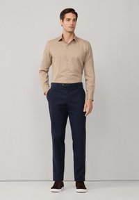 Beige overhemd met lange mouwen en knopen, gecombineerd met marineblauwe broek. Het model draagt bruine sneakers met witte zolen. Gewerkt textiel zichtbaar.