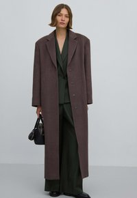 Lang bruin wollen coat met een brede revers en vijf knopen, gedragen over een donkergroene pak. Zwarte handtas met accenten vastgehouden in één hand.
