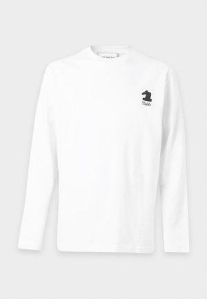 Uomo con camicia bianca a maniche lunghe con piccolo logo nero del cavaliere, jeans chiari e larghi e scarpe nere in piedi su sfondo bianco.