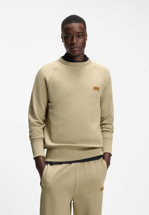 Homme portant un sweat-shirt beige et un pantalon assorti, mains dans les poches, sur fond blanc uni.