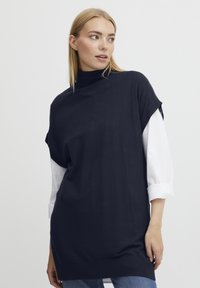 Tunic a maglia blu navy con alta scollatura, maniche corte e spacchi laterali; indossata sopra una camicia bianca con maniche arrotolate.